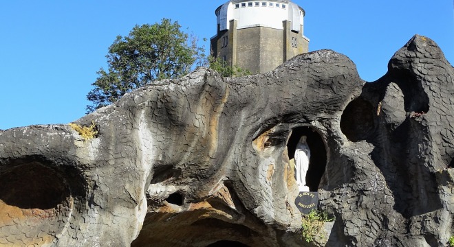 Beeld van een Mariabeeld in een grot met op de achtergrond een toren en een blauwe lucht.