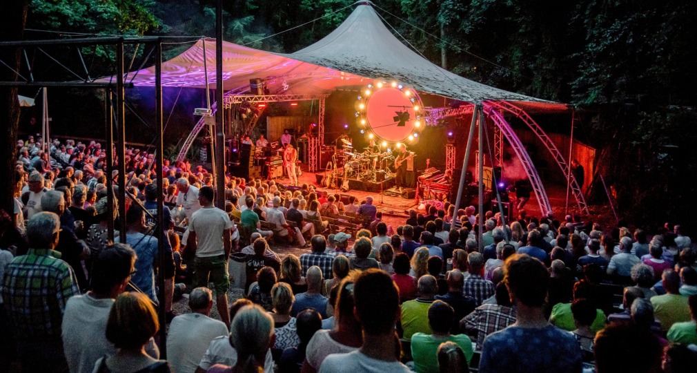 Publiek zit buiten in een amfitheater om te genieten van een live concert onder een verlichte tent.