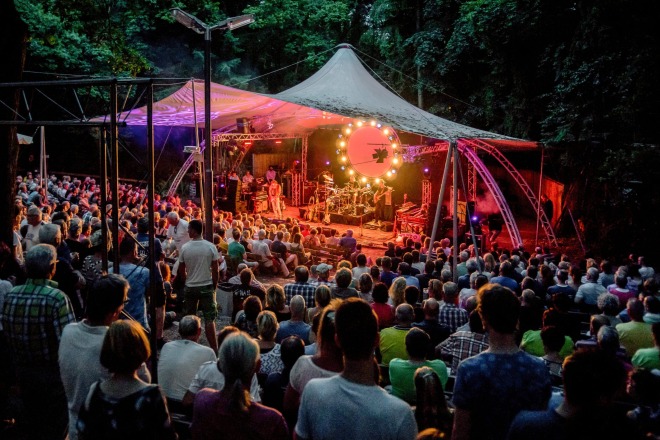 Publiek zit buiten in een amfitheater om te genieten van een live concert onder een verlichte tent.