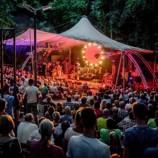 Publiek zit buiten in een amfitheater om te genieten van een live concert onder een verlichte tent.