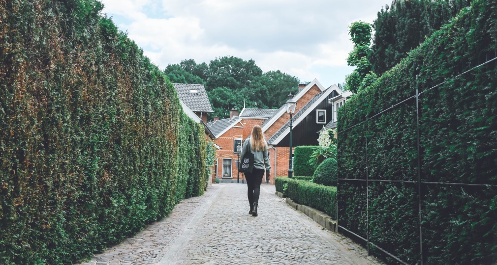 Vrouw wandelt over een smalle, geplaveide straat omringd door hoge heggen en traditionele huizen.