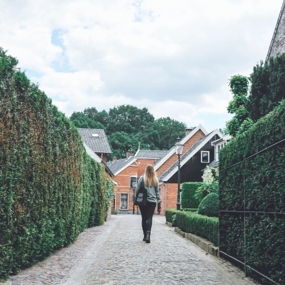 Vrouw wandelt over een smalle, geplaveide straat omringd door hoge heggen en traditionele huizen.