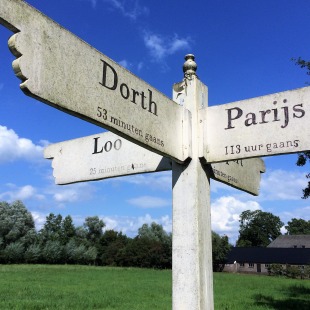 Wegwijzer op het platteland met richtingen naar Dorth, Parijs en Loo, onder een heldere blauwe lucht.