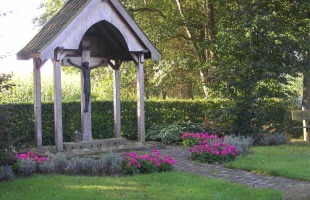 Buitenbeeld van een houten kruisbeeld met Christus onder een afdak, omringd door bloemen en groen gras.