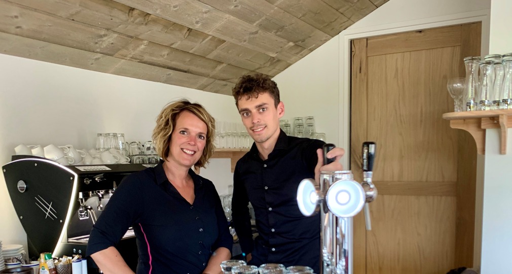 Zwei Personen stehen hinter einer Bar in einem Café mit Gläsern, Zapfanlage und Espressomaschine.