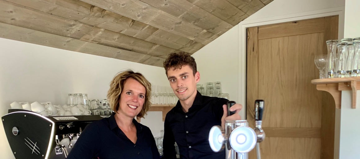 Twee mensen achter een bar in een café met glazen, een biertap en een espressomachine op de achtergrond.