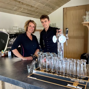 Twee mensen achter een bar in een café met glazen, een biertap en een espressomachine op de achtergrond.