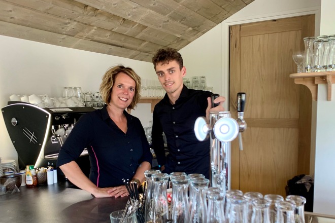 Twee mensen achter een bar in een café met glazen, een biertap en een espressomachine op de achtergrond.