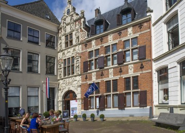 Buitenaanzicht van een historisch gebouw met terras en mensen die genieten op een zonnige dag in Nederland.