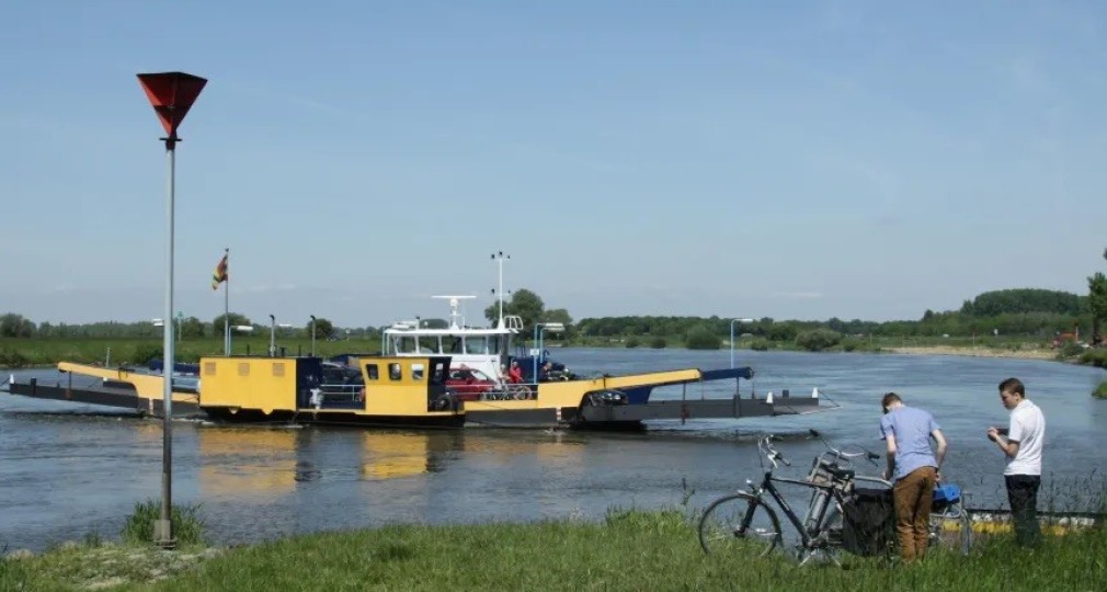 Twee mannen met fietsen wachten bij het water terwijl een gele veerpont voertuigen over de rivier vervoert.