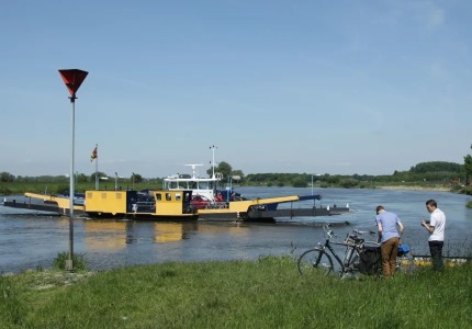 Twee mannen met fietsen wachten bij het water terwijl een gele veerpont voertuigen over de rivier vervoert.
