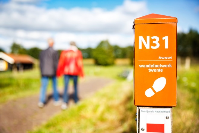 Oranje wandelroutepaal met opschrift N31 en wandelnetwerk Twente, twee wandelaars op de achtergrond