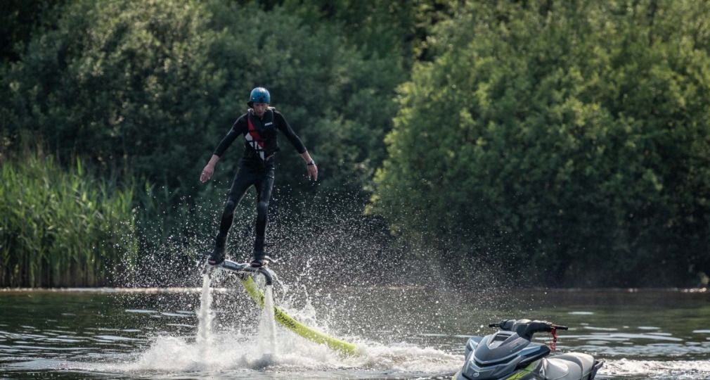 Persoon met helm en wetsuit vliegt boven water op een flyboard, met jetski en bomen op de achtergrond.