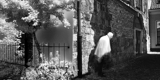 Zwart-wit foto van een onscherpe persoon die langs een stenen muur loopt, met een heldere boom links.