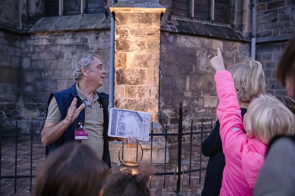 Een gids met een boek leidt een groep mensen tijdens een avondwandeling bij een oud stenen gebouw.