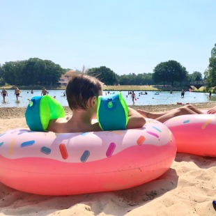 Twee kinderen ontspannen op roze donut-zwembanden op een zonnig strand met meer op de achtergrond.