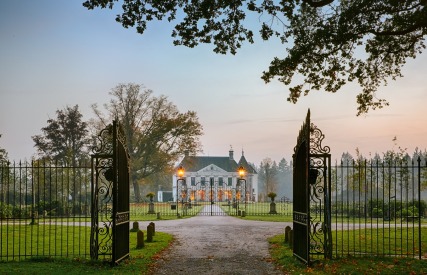 Toegang tot een majestueus landhuis met smeedijzeren poort, verlichte lantaarns en ruime tuin bij zonsopkomst.