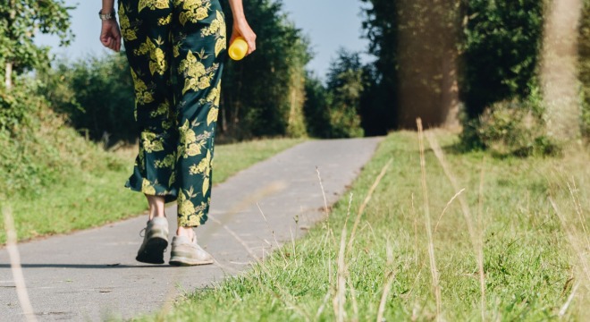 Persoon wandelt op een landelijk pad, draagt een gele trui en gebloemde broek, flesje in hand.