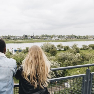 Twee mensen kijken uit over een rivierlandschap met groene bomen, wandelpaden en een windmolen.