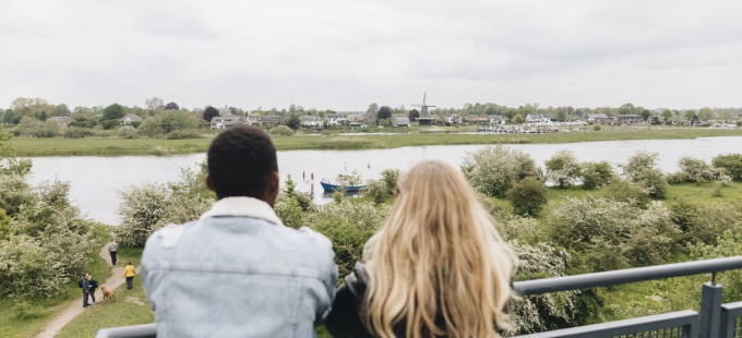 Twee mensen kijken uit over een rivierlandschap met groene bomen, wandelpaden en een windmolen.
