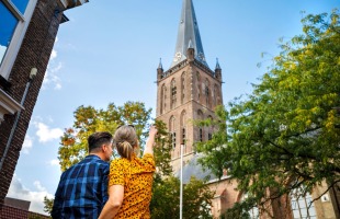 Stel kijkt omhoog naar een historische kerktoren omringd door bomen en blauwe lucht op een zonnige dag.