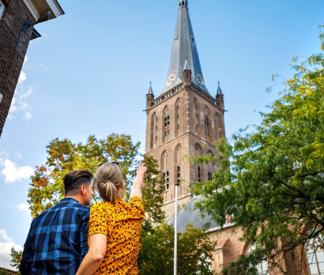 Stel kijkt omhoog naar een historische kerktoren omringd door bomen en blauwe lucht op een zonnige dag.