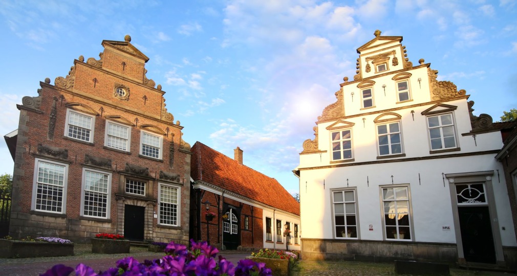 Oude Nederlandse herenhuizen met trapgevels en bloembedden op een geplaveid dorpsplein bij zonsondergang.