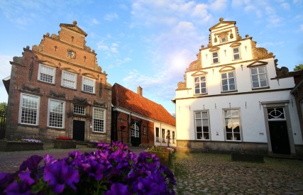 Oude Nederlandse herenhuizen met trapgevels en bloembedden op een geplaveid dorpsplein bij zonsondergang.