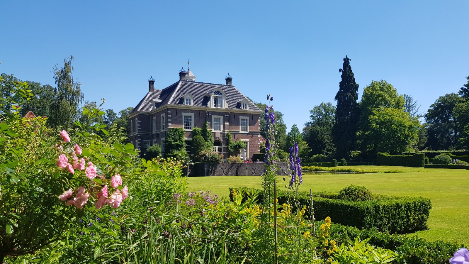 Elegant landhuis met statige tuin vol bloemen en struiken onder een heldere blauwe lucht in de zomer.
