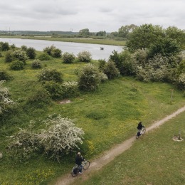 Twee mensen fietsen over een smal pad langs groene struiken en een rivier onder een bewolkte lucht.