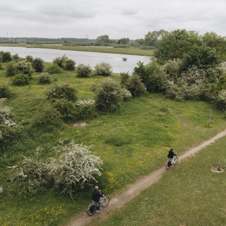 Twee mensen fietsen over een smal pad langs groene struiken en een rivier onder een bewolkte lucht.