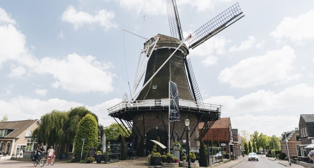 Traditionele Nederlandse windmolen in een schilderachtig dorp met mensen op fietsen en blauwe lucht.