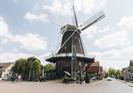 Traditionele Nederlandse windmolen in een schilderachtig dorp met mensen op fietsen en blauwe lucht.