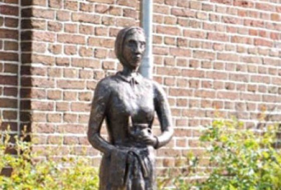 Bronzen standbeeld van een vrouw in traditionele kleding op een sokkel, buiten naast een bakstenen muur.