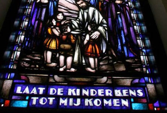 Gebrandschilderd raam met Jezus die kinderen zegent en de tekst: LAAT DE KINDERKENS TOT MIJ KOMEN.