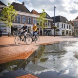 Twee mensen fietsen langs een fontein op een zonnig plein met traditionele huizen in Nederland.
