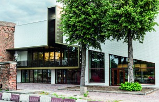 Moderne witte gevel van een gebouw met grote ramen, twee bomen ervoor en een bakstenen muur links.