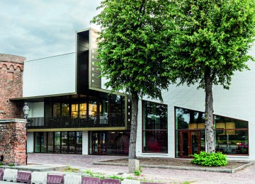 Moderne witte gevel van een gebouw met grote ramen, twee bomen ervoor en een bakstenen muur links.