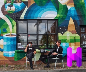Twee mensen zitten buiten voor een kleurrijke muurschildering met een kat en vrouw, drinken koffie.