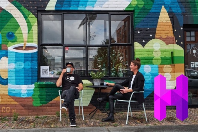 Twee mensen zitten buiten voor een kleurrijke muurschildering met een kat en vrouw, drinken koffie.