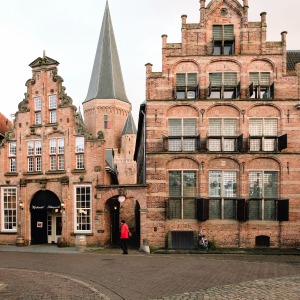 Historische bakstenen gebouwen met trapgevels en toren in een Nederlandse stad, persoon in rood jack loopt.