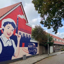 Muurschildering van een vrouw met koekje op een bakstenen huis, rustige straat met bomen en auto’s.