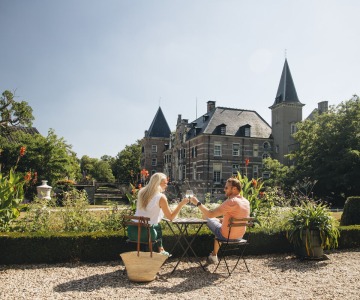 Twee mensen genieten van een drankje aan een tafel in een tuin bij een kasteel op een zonnige dag.