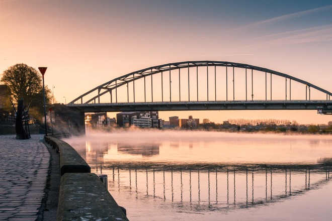Een elegante boogbrug over een rustige rivier bij zonsopgang, met reflecties en bomen aan de oever.