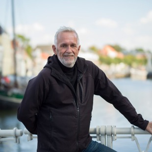 Oudere man met grijs haar en baard leunt tegen een brugleuning met water en huizen op de achtergrond.