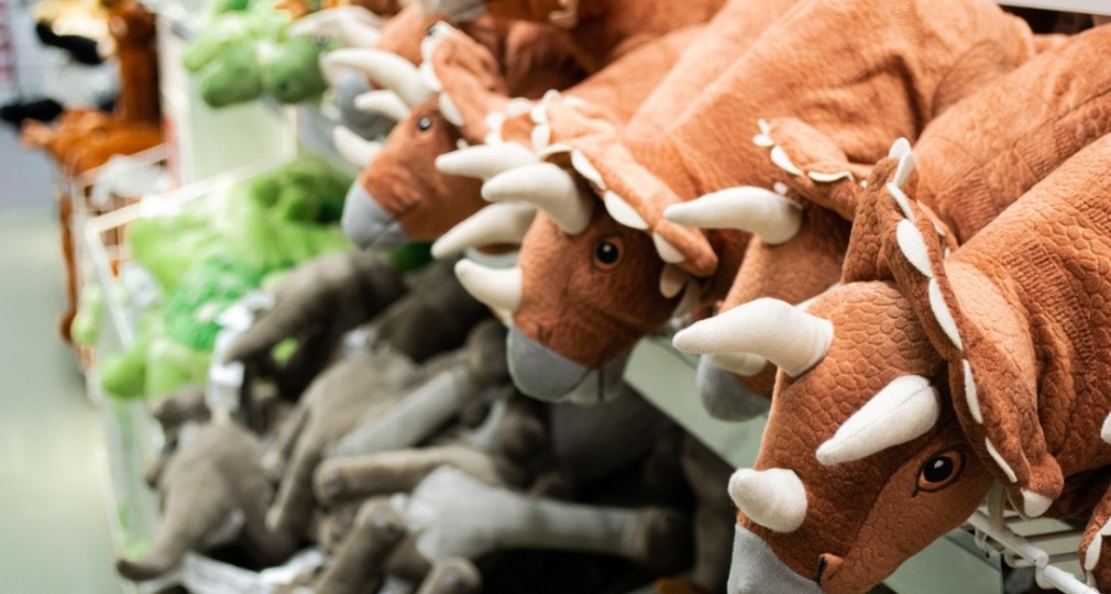 Knuffeldieren in de vorm van dinosaurussen liggen opgestapeld op planken in een winkel of speelgoedafdeling.