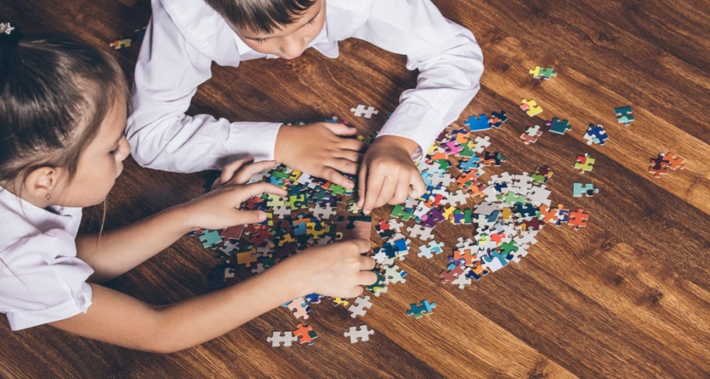 Twee kinderen liggen op een houten vloer en werken samen aan een kleurrijke puzzel met losse stukjes.