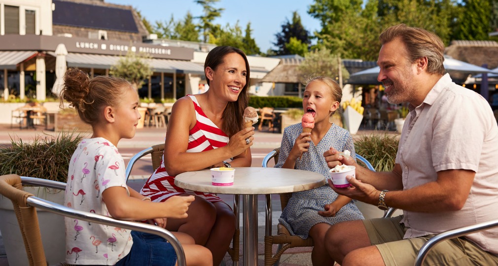 Gezin met twee kinderen eet samen ijsjes aan een buitentafel bij een restaurant op een zonnige dag.
