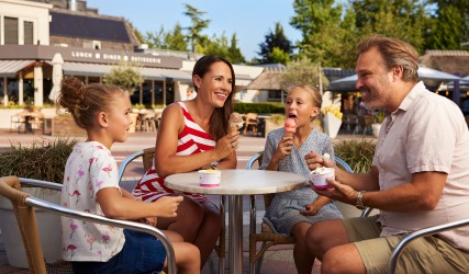 Gezin met twee kinderen eet samen ijsjes aan een buitentafel bij een restaurant op een zonnige dag.