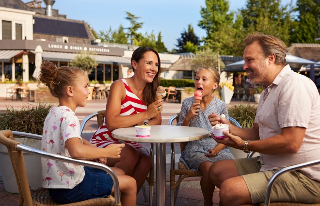 Gezin met twee kinderen eet samen ijsjes aan een buitentafel bij een restaurant op een zonnige dag.